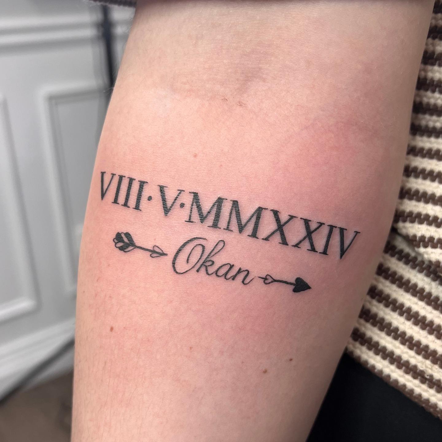 Fine line script tattoo with a name, Roman numeral date and heart arrow by Independence Ink.